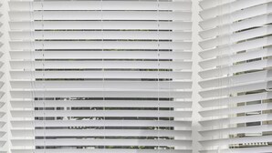 Una silla con patas de madera y asiento tapizado en gris frente a una ventana con persianas blancas.