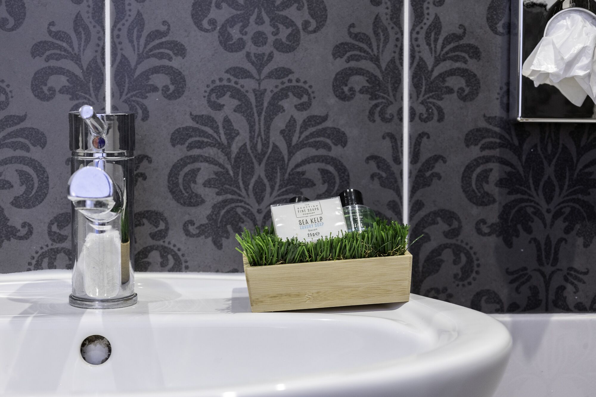 A bathroom sink with a clear glass soap dispenser, a wooden soap dish with a bottle of Sea Kelp Luxury Soap, and a tissue box on the wall.