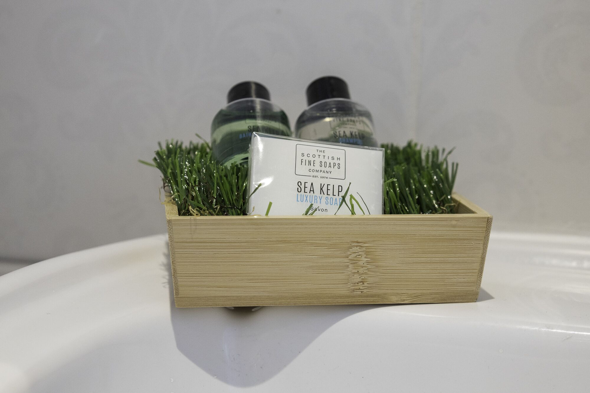 Una caja de madera con dos botellas de Jabón de Algas Marinas de Lujo y una etiqueta, sobre una superficie blanca con adornos que simulan hierba verde.