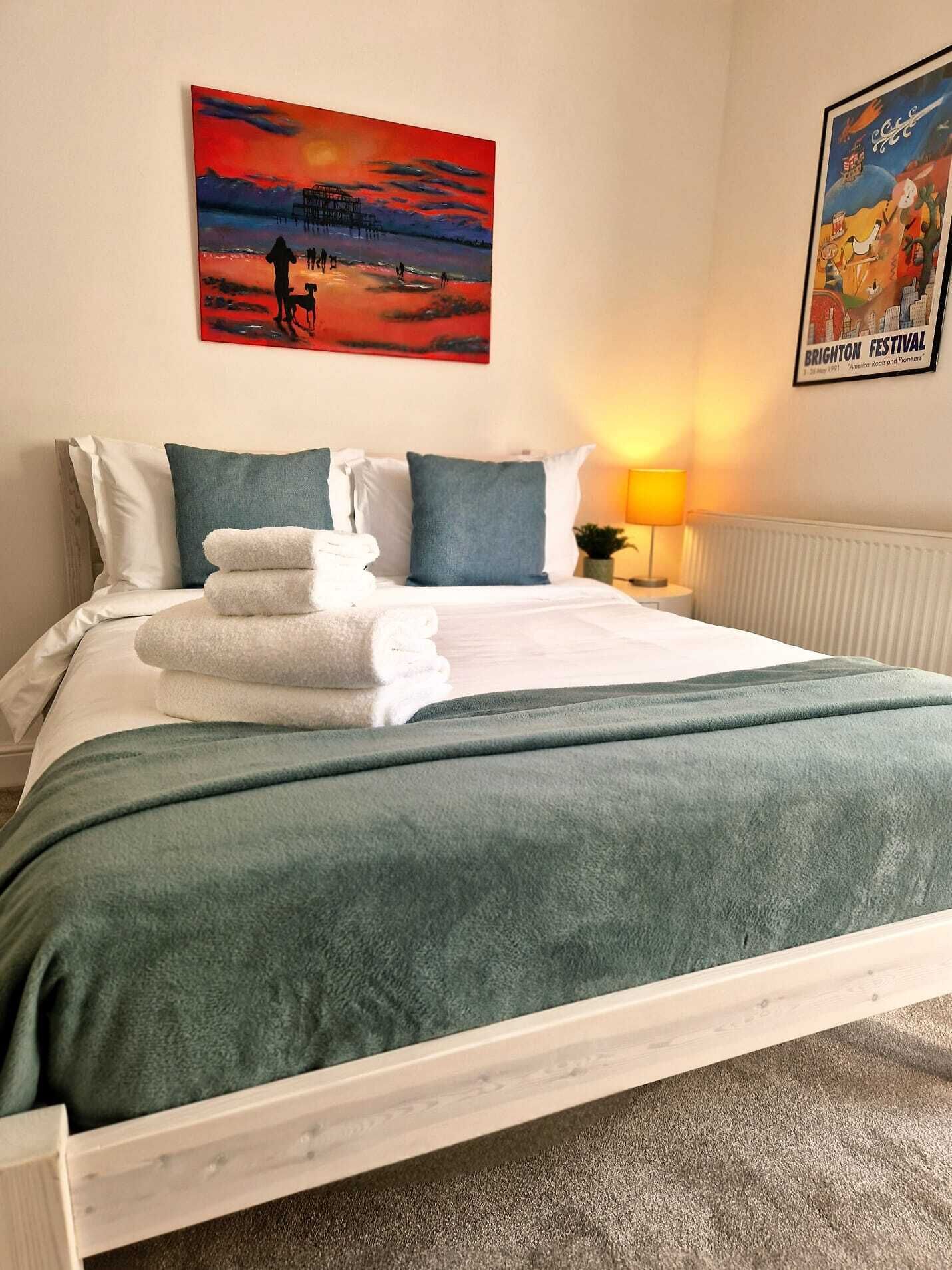 Ein Hotelzimmer mit Bett, Kissen, Decke, einem Gemälde, einer Lampe und einem Poster an der Wand.