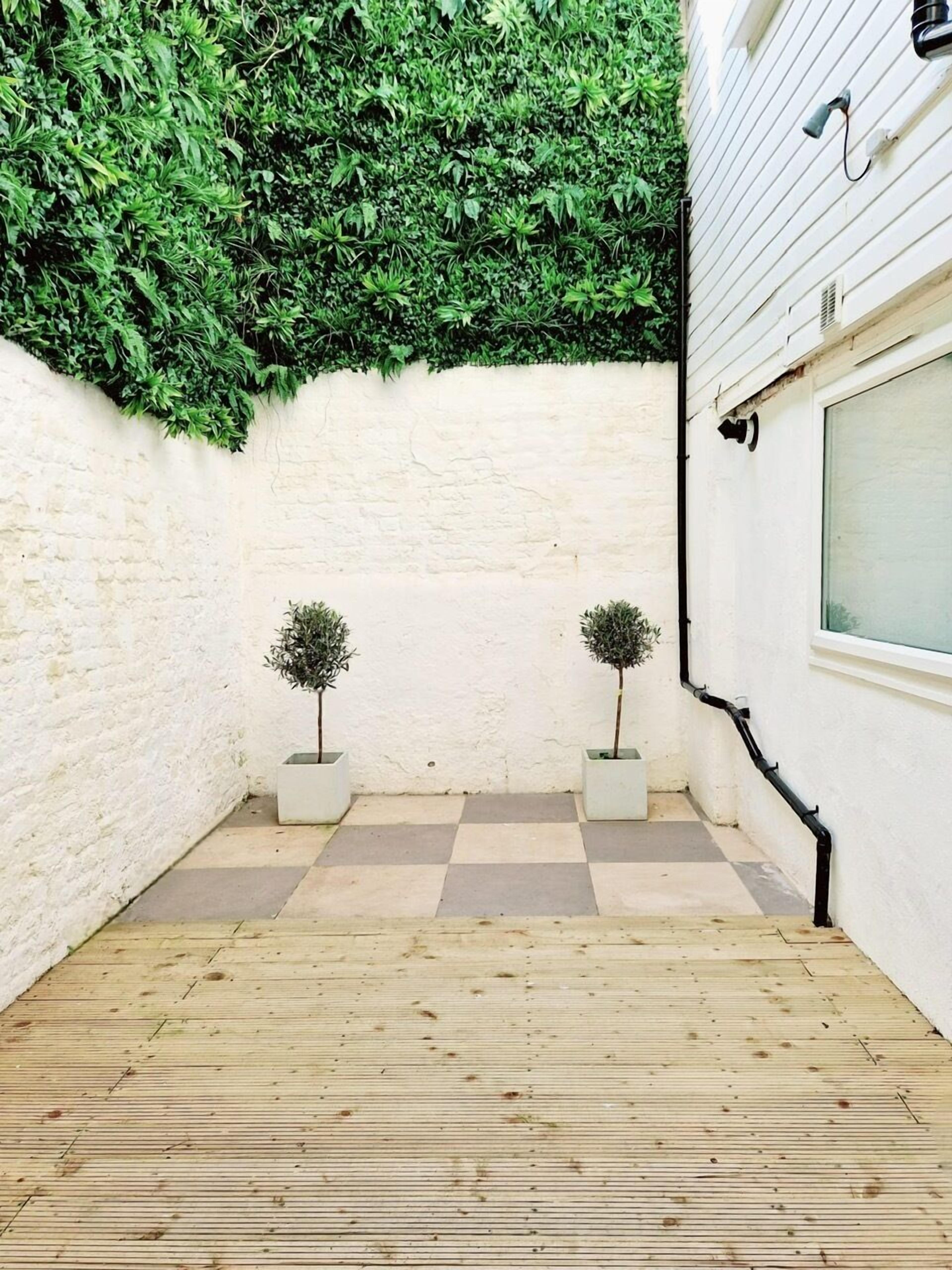 A small, enclosed outdoor space with a checkered floor, two potted trees, and a white wall with a green leafy wall behind it.