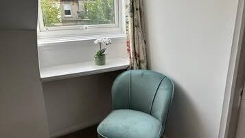 Una habitación de esquina con una silla color turquesa, una ventana con una orquídea en maceta y un cuadro en la pared.