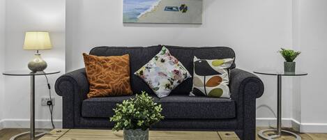 A living room with a dark grey sofa, three cushions, a small round table with a lamp, a potted plant, and a painting of a beach scene on the wall.