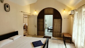 A hotel room with a bed, a small table, a chair, and a view through an arched doorway. - NuaGoa Nature Retreat Private Limited (Cuttack)