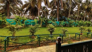 A lush garden with palm trees and banana plants, a thatched-roof structure, and a green metal railing. - NuaGoa Nature Retreat Private Limited (Cuttack)