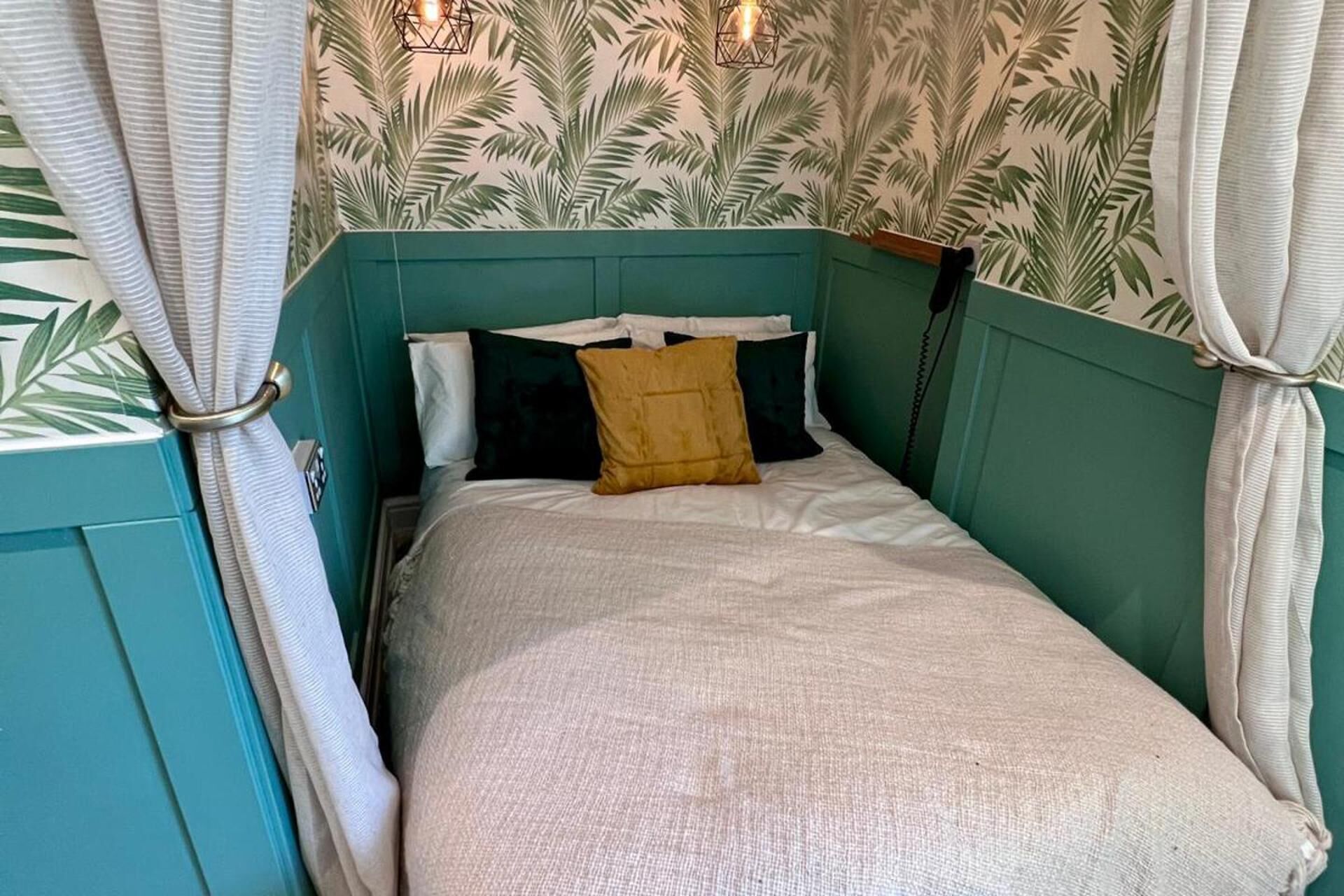 A single bed with a white bedspread and a mustard-colored pillow, set against a teal headboard and walls with a palm leaf pattern.