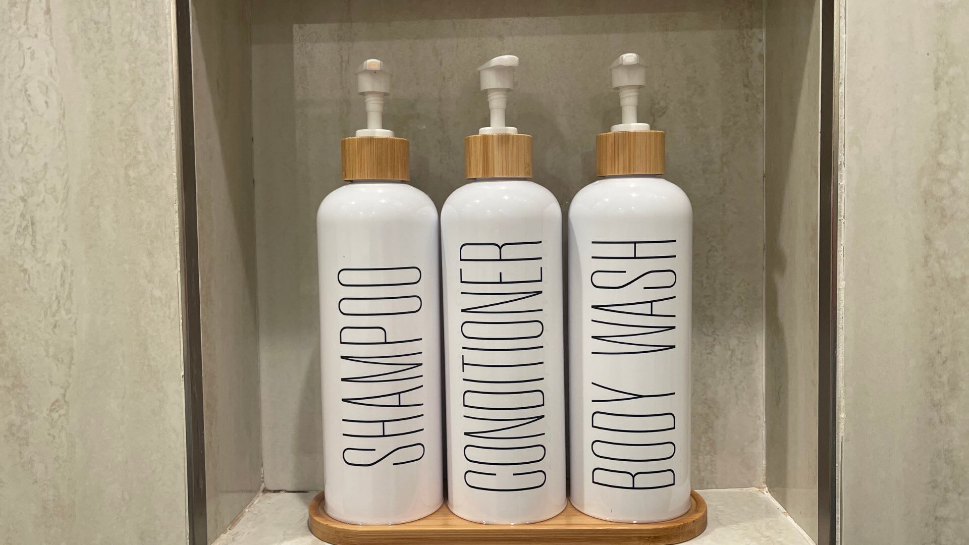 Three white bottles with wooden caps labeled "SHAMPOO," "CONDITIONER," and "BODY WASH" on a wooden stand.