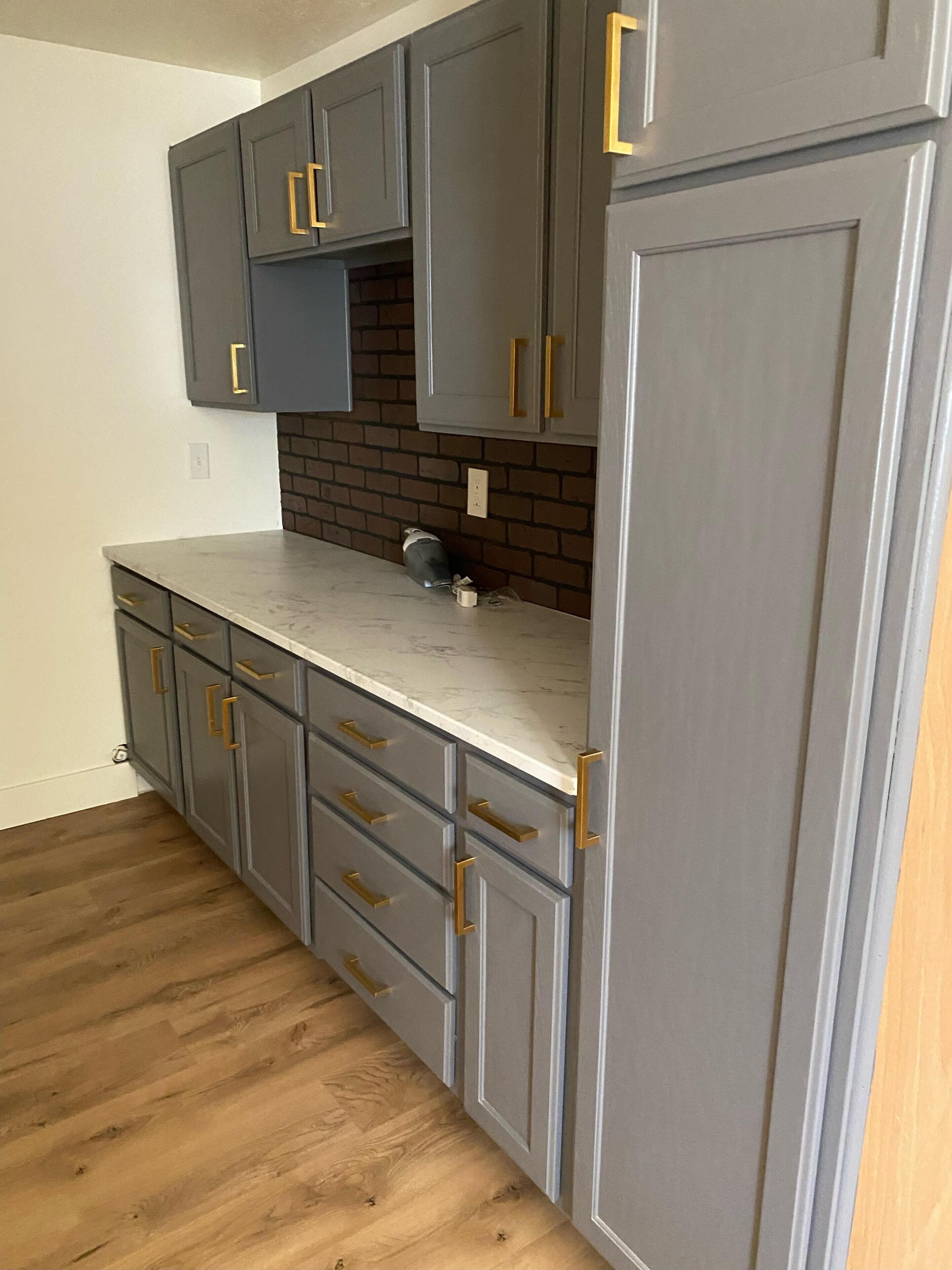 A kitchen with grey cabinets, a marble countertop, and a brick backsplash.