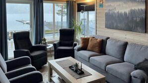 En moderne stue med sofa, lænestole og sofabord. Store vinduer giver udsigt over vand og en bro.