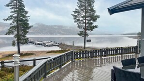 En træterrasse med gelænder, der kigger ud over en rolig sø, omgivet af træer og fjerne bjerge.