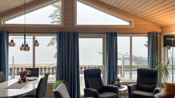 En rummelig stue med store vinduer, træloft og et spiseområde med bord og stole.