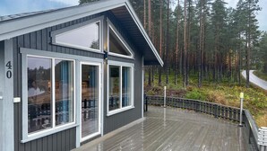 En moderne hytte med store vinduer, en terrasse og skov i baggrunden.