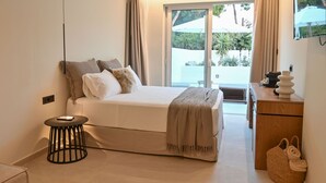Ein Schlafzimmer mit einem Bett, einem kleinen runden Tisch, einer hölzernen Kommode und Blick ins Freie durch eine Schiebetür.