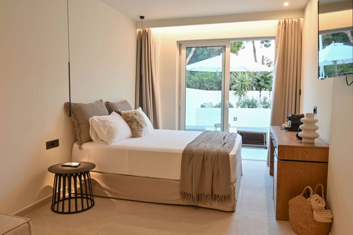 A bedroom with a bed, a small round table, a wooden dresser, and a view of the outdoors through a sliding door.