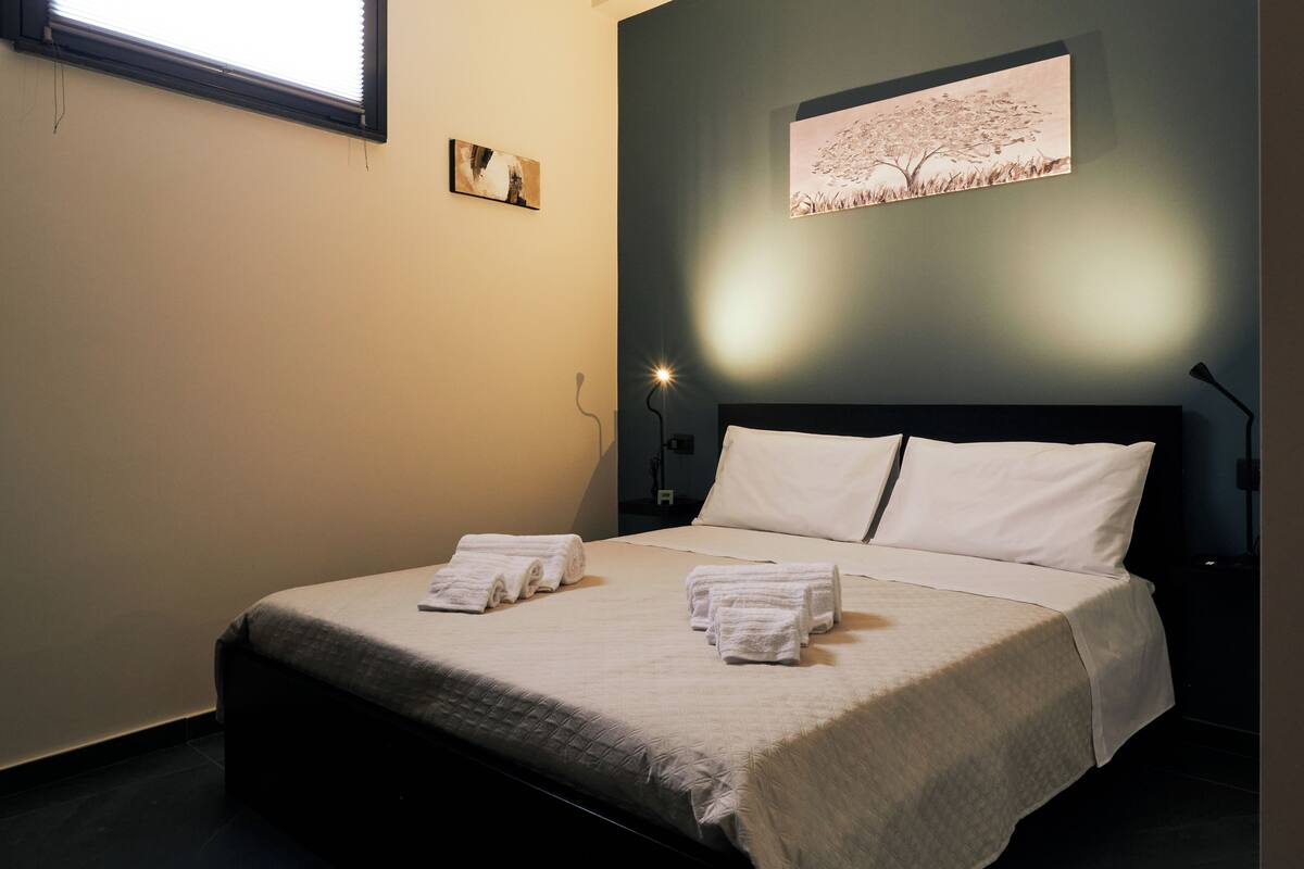 A neatly made bed with white linens and towels, a dark headboard, and a wall-mounted artwork of a tree.