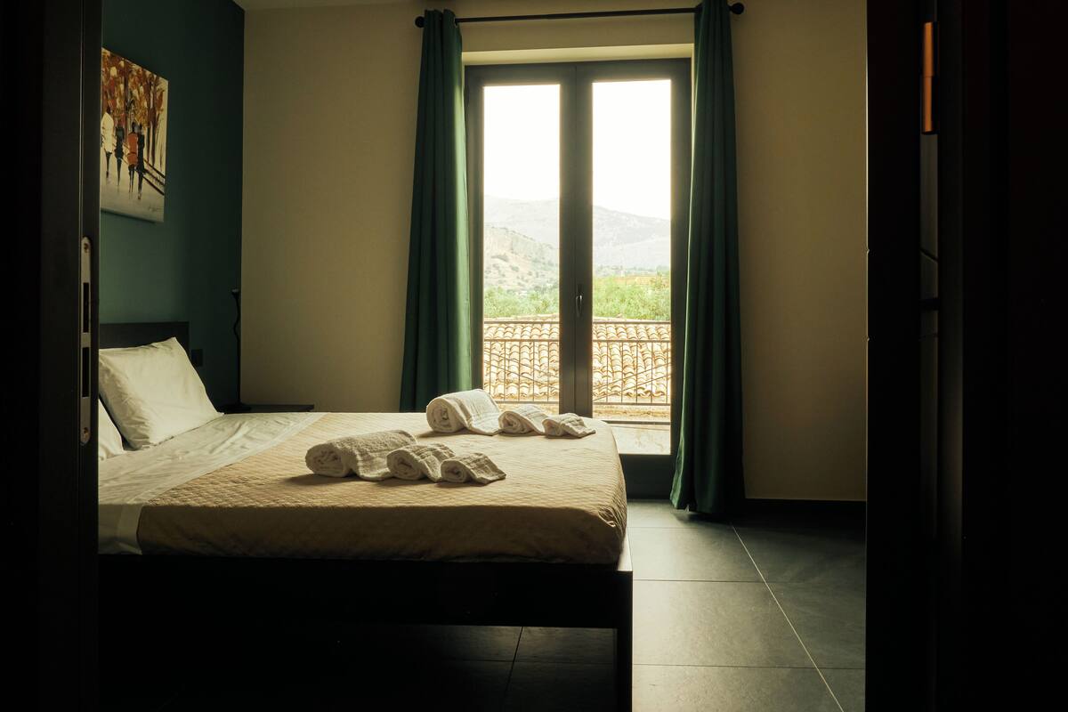 A hotel room with a bed, a view of the outdoors through a window, and a painting on the wall.
