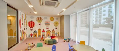 A brightly lit children's playroom with colorful chairs and tables, a large window, and playful wall decorations.