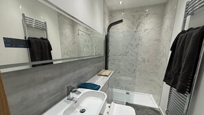 A modern bathroom with a white sink, a toilet, and a shower with a glass door. There is a towel rack on the wall and a small shelf with towels. - Exquisite Riverside Living in a Stunning Flat (Gravesend)
