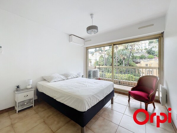 A bedroom with a bed, a nightstand, a chair, and a large window with a view of buildings and trees.