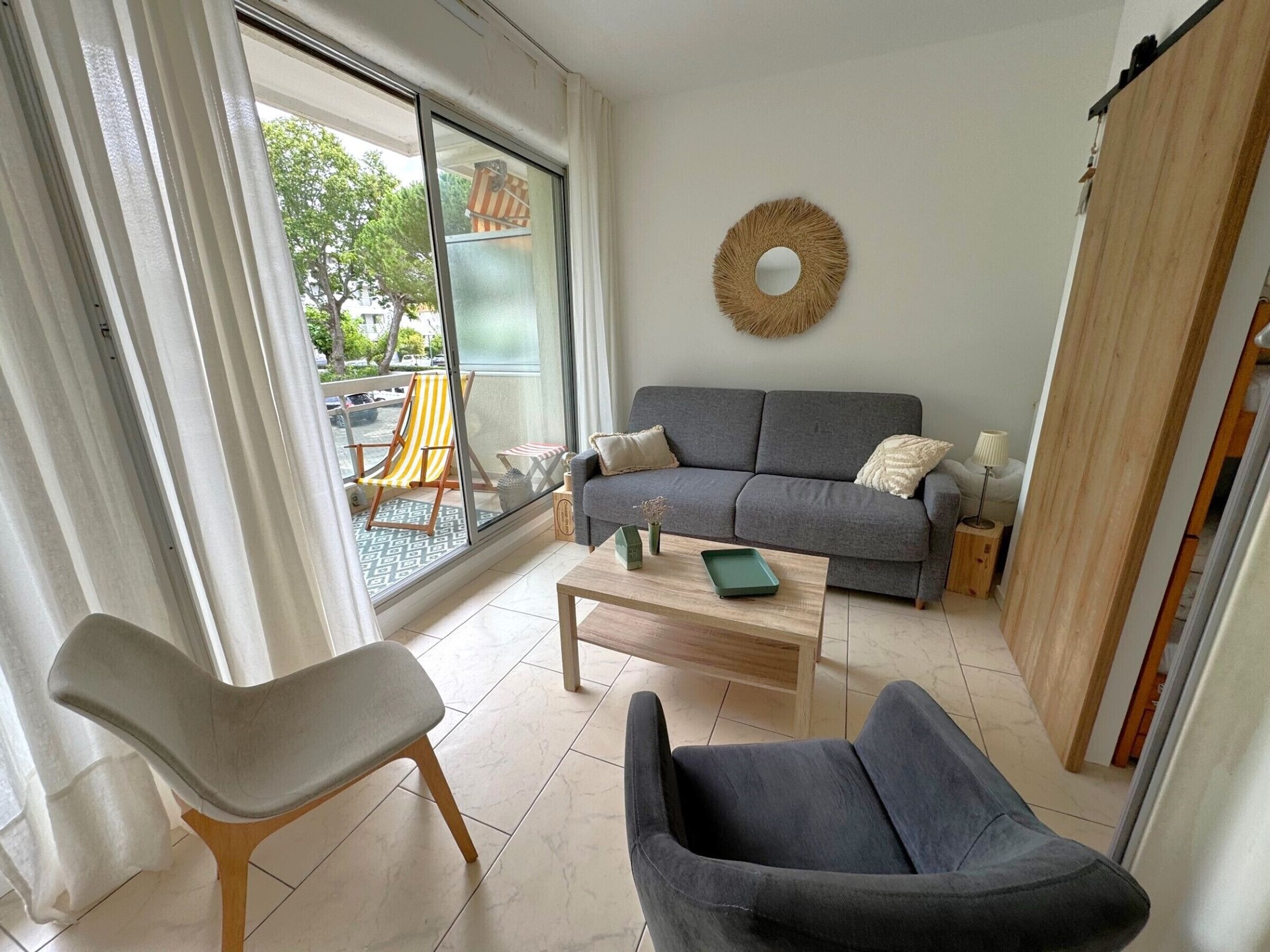Un salon moderne avec un canapé gris, une table basse en bois, un fauteuil gris et une chaise à rayures placée à l’extérieur de la porte-fenêtre coulissante.