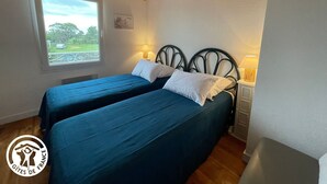 A room with two beds, a window with a view of greenery, and a wooden ceiling.