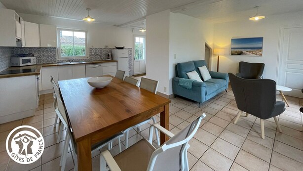 A modern kitchen with white cabinets and a wooden dining table. There is a blue sofa and a black armchair in the living area.