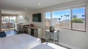 A hotel room with a bed, a desk, a chair, a TV, and a balcony with a view of palm trees and the beach. - Ocean-View Home Blocks to Beach with Private Hot Tub & Rooftop Deck! (San Diego)