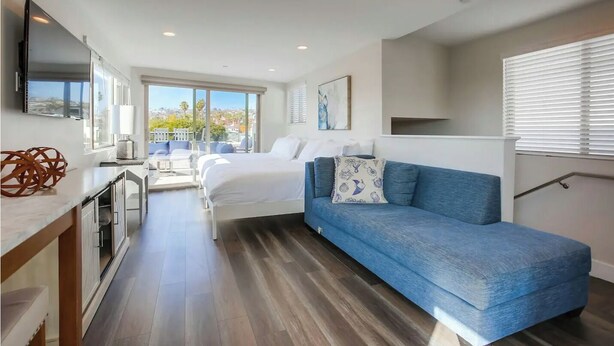 A modern hotel room with a bed, a blue sofa, a small kitchen area, and a balcony with a view. - Ocean-View Home Blocks to Beach with Private Hot Tub & Rooftop Deck! (San Diego)