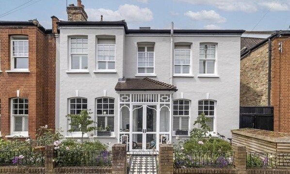 A two-story house with a white facade, multiple windows with shutters, and a small front garden with a tiled entrance.