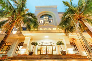 El exterior del New Ha Long Hotel, con una entrada imponente, palmeras y un cielo despejado.