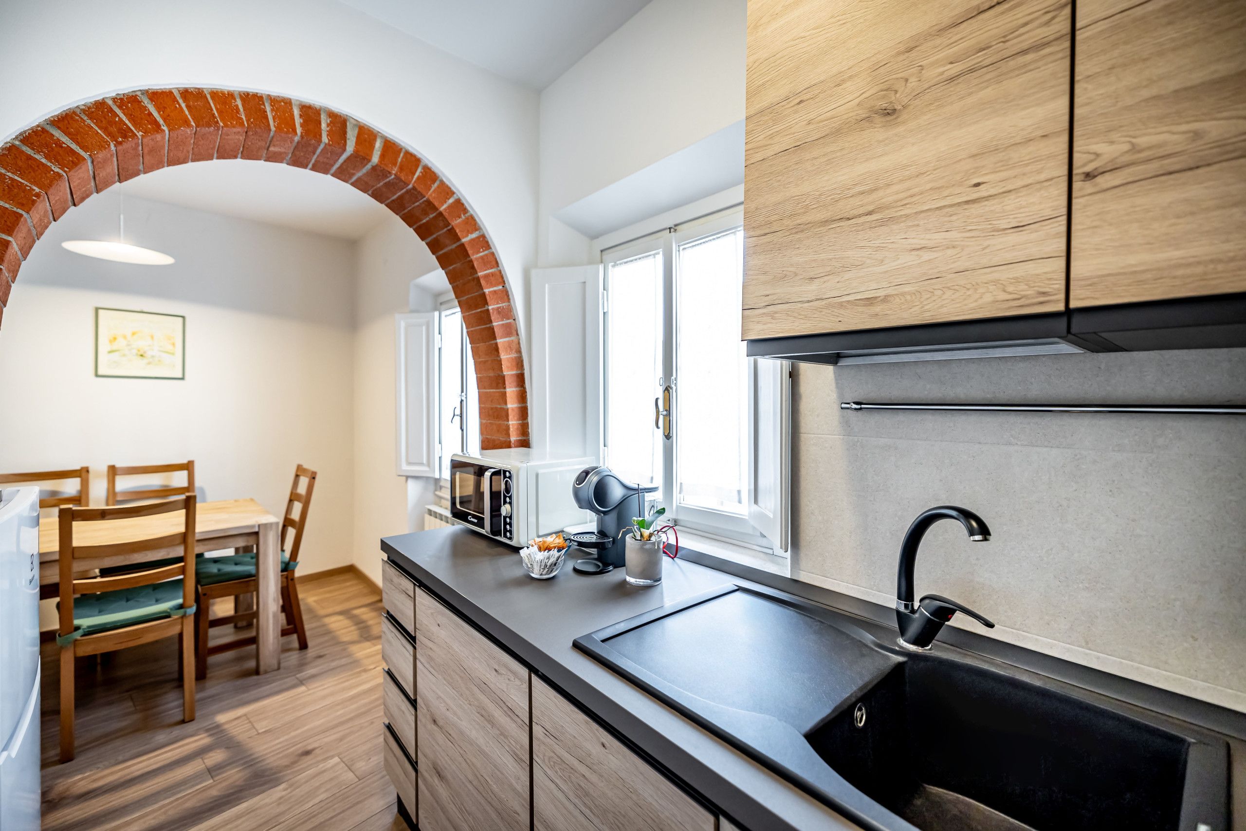 A kitchen with a brick archway, wooden cabinets, a black sink, and a dining area with chairs and a table.