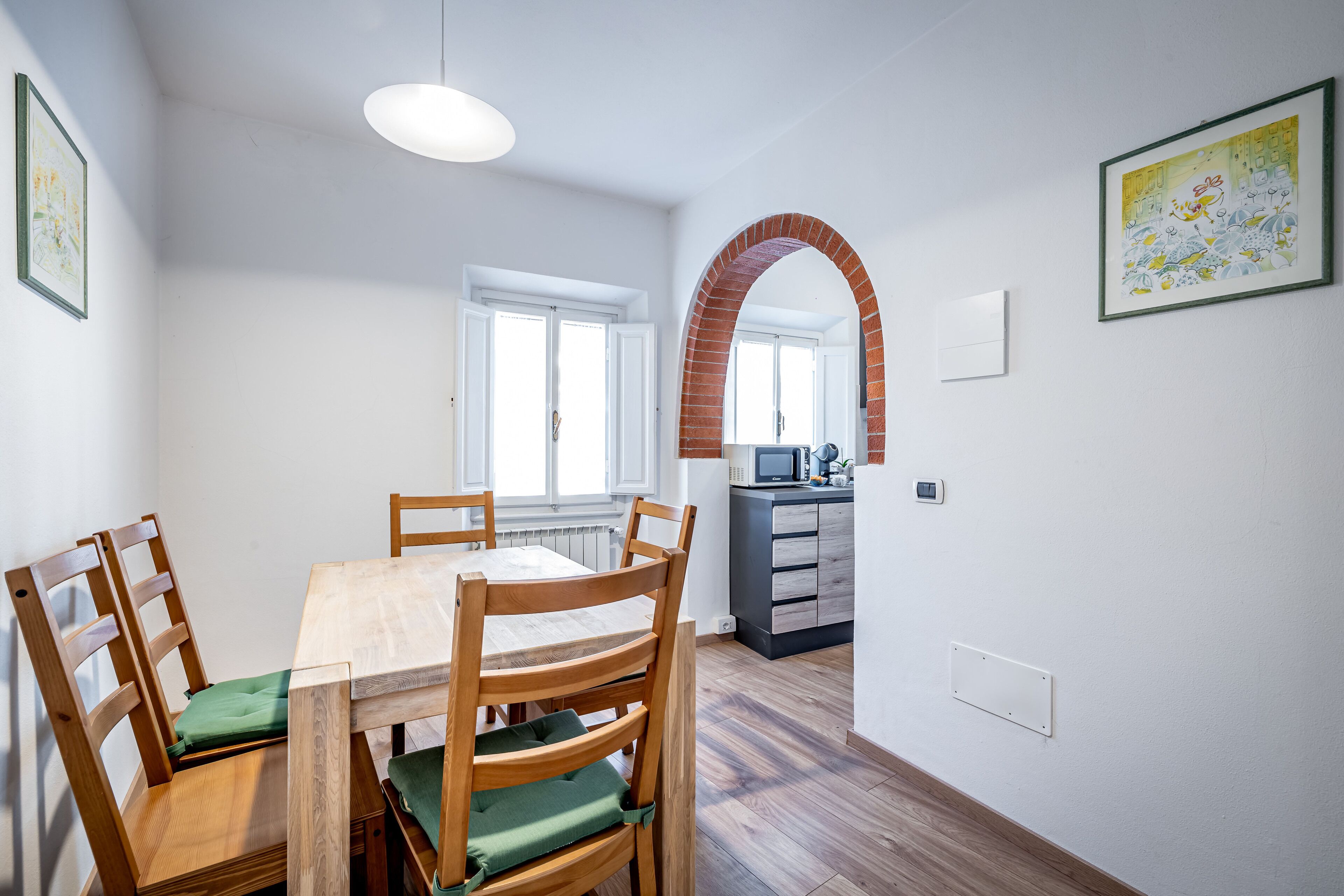 A dining area with a wooden table and chairs, a kitchen in the background, and a brick archway.
