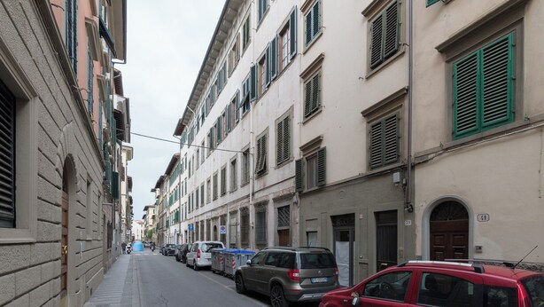A narrow street lined with multi-story buildings, parked cars, and a sidewalk. - YiD Pilastri (Florence)