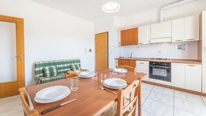 A dining area with a wooden table set for four, a green plaid sofa, and a kitchen with white cabinets and a built-in oven. - Apartment Balcony Sea View - Gallipoli (Gallipoli)