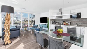 Um apartamento moderno com sala de estar, incluindo sofá, cadeiras e mesa de jantar de vidro. Há uma cozinha com armários brancos, micro-ondas Bosch e uma janela grande com vista para a cidade.