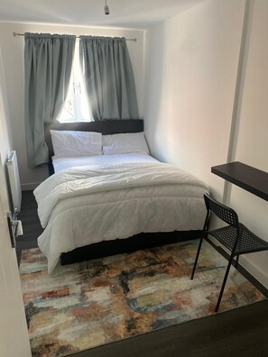 Un dormitorio con cama, una silla, un escritorio y una ventana con cortinas.