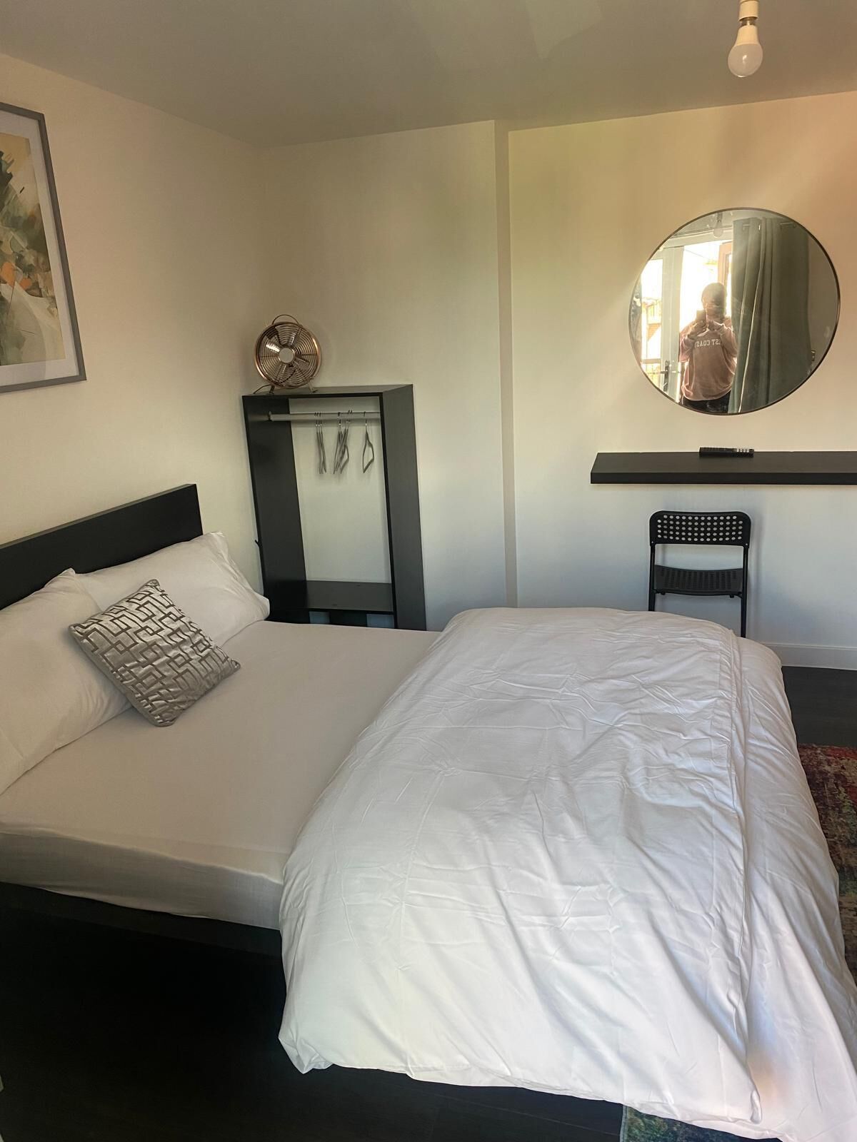 A neatly made bed with a white comforter and a patterned pillow, a round mirror on the wall, and a small chair.