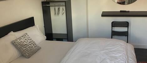 A neatly made bed with a white comforter and a patterned pillow, a round mirror on the wall, and a small chair.
