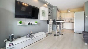 A modern living room with a flat-screen TV, a white console table, and a dining area with two bar stools. - RentPlanet - Pogodne Osiedle (Kolobrzeg)