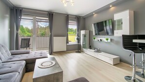 A modern living room with a grey sectional sofa, a coffee table, a TV mounted on the wall, and a large window with curtains. - RentPlanet - Pogodne Osiedle (Kolobrzeg)