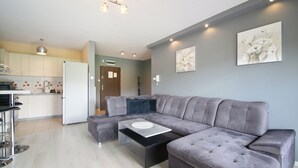 A modern living room with a grey sectional sofa, a coffee table, and a kitchen area with white cabinets and a refrigerator. - RentPlanet - Pogodne Osiedle (Kolobrzeg)