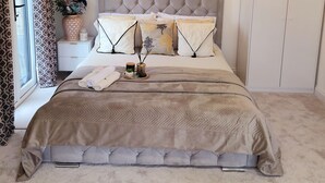 Une chambre à coucher avec une tête de lit capitonnée, une table de chevet ornée d’un vase et de bougies, un grand lit recouvert d’une couverture beige et une armoire.