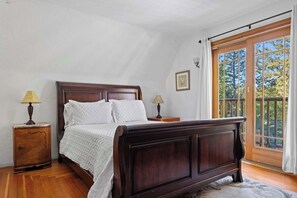Un dormitorio con una cama de madera, dos mesitas de noche con lámparas, un ventanal con vistas a árboles y un cuadro en la pared.