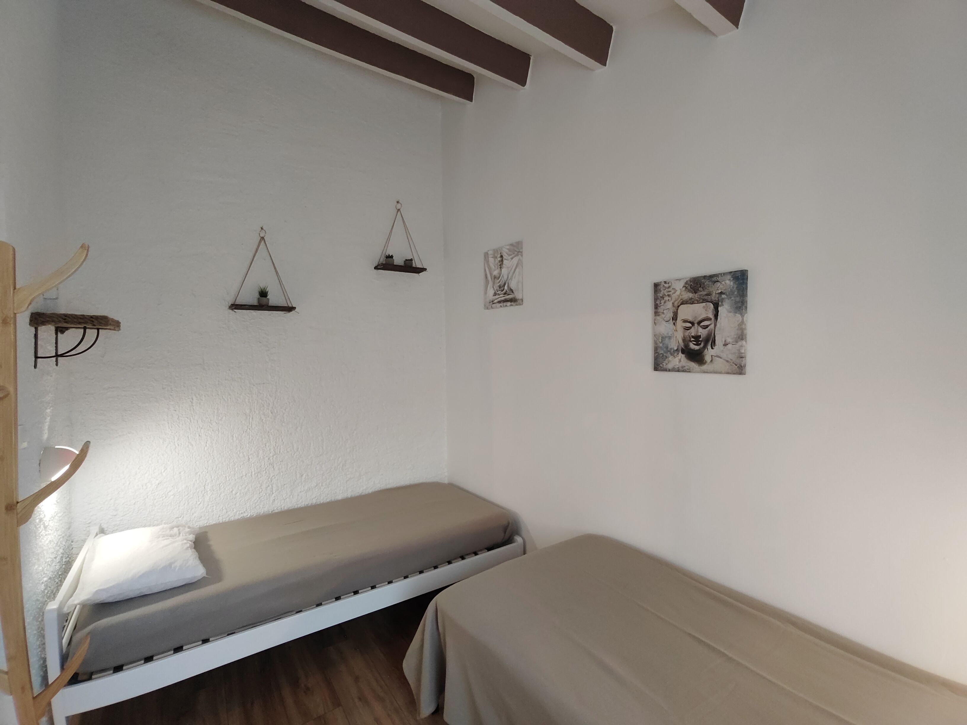 A small, single-bed room with a wooden bench, a wall-mounted shelf with a plant, and a Buddha statue.