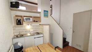 Une cuisine compacte équipée d’un four à micro-ondes, d’une machine à café et d’un évier. On y trouve une table en bois et une poubelle. Un escalier mène à une porte.