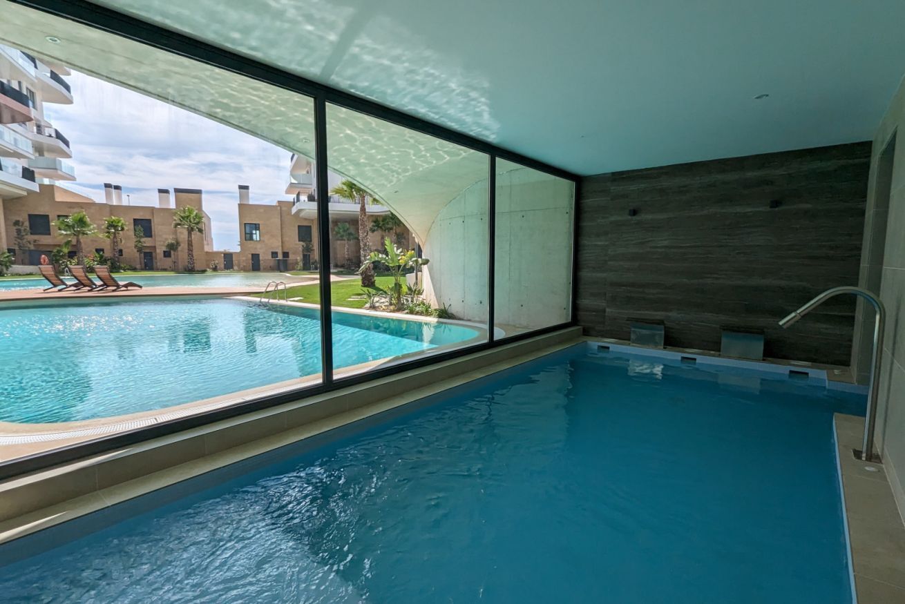An indoor swimming pool with clear views of a poolside area, including lounge chairs and a building with balconies in the background.