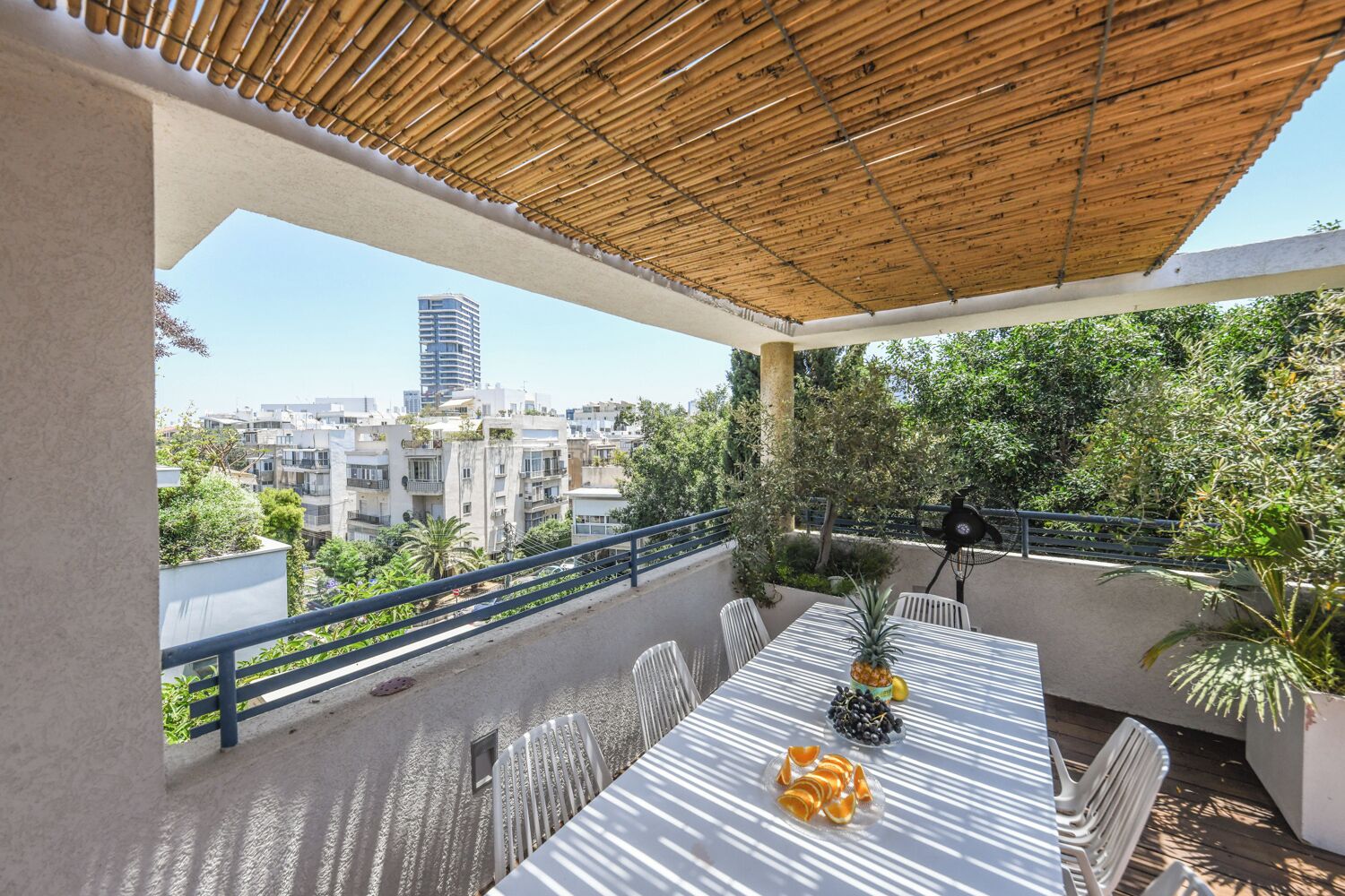 A balcony with a table, chairs, and a fan. There is a view of buildings and trees in the background.