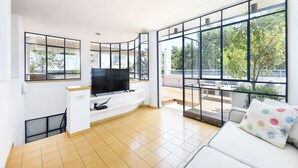 A modern living room with a flat-screen TV on a white entertainment unit, large windows offering a view of the outdoors, and a white sofa with a colorful pillow. - Spacious 3BR Penthouse w Rooftop (Tel Aviv)