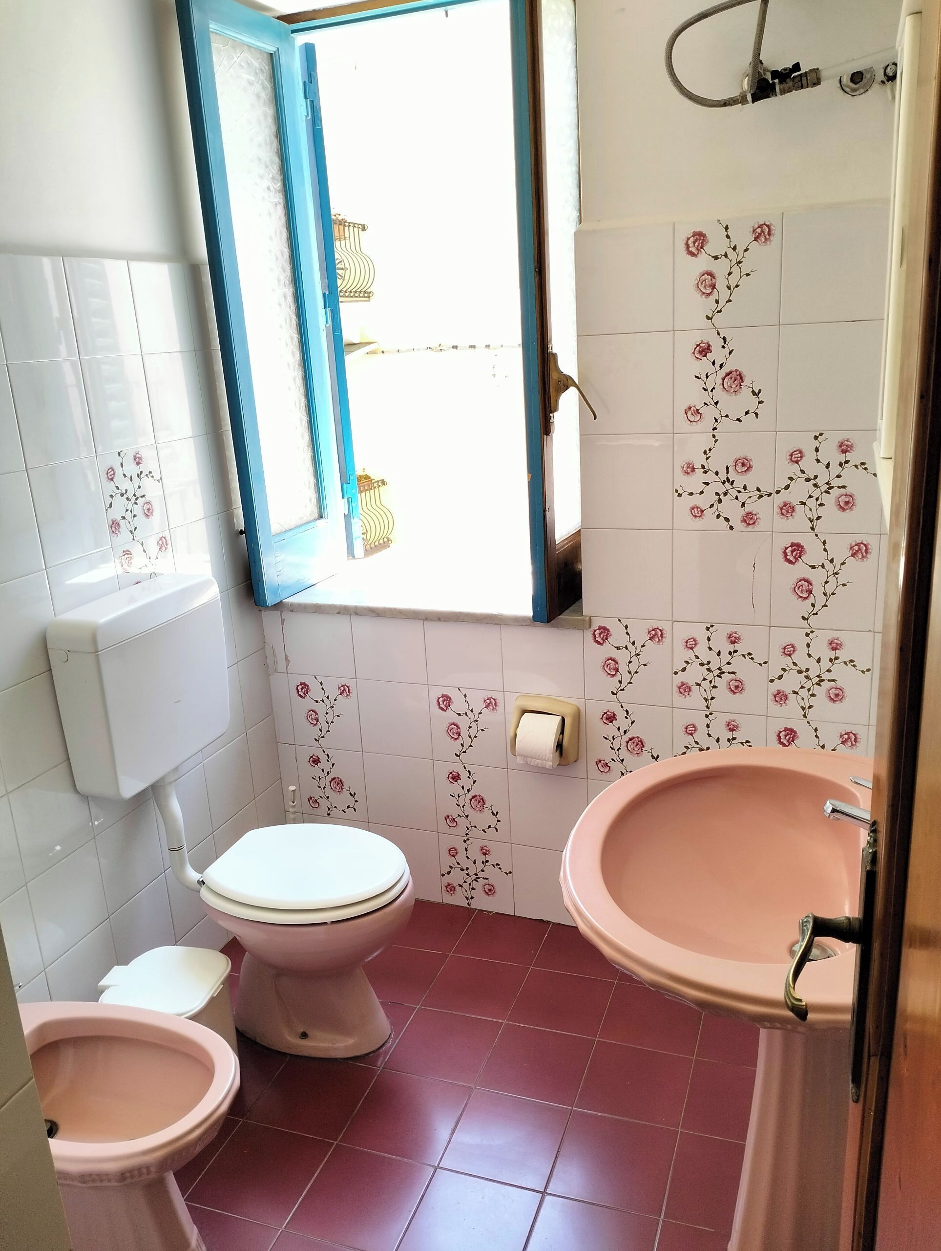 Baño con inodoro, bidé y lavamanos, con azulejos de motivos florales y una ventana con marco azul.