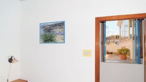 Une chambre à coucher comprenant un lit, une table de chevet, une lampe fixée au mur, un tableau et une fenêtre donnant sur des immeubles et une rue.